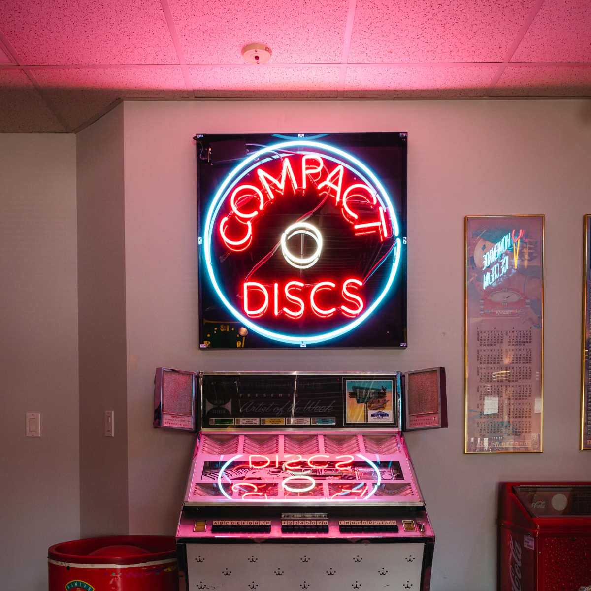 A vintage jukebox featuring a neon sign that reads "COMPACT DISCS" stands against a wall in a room with black and white checkered flooring.