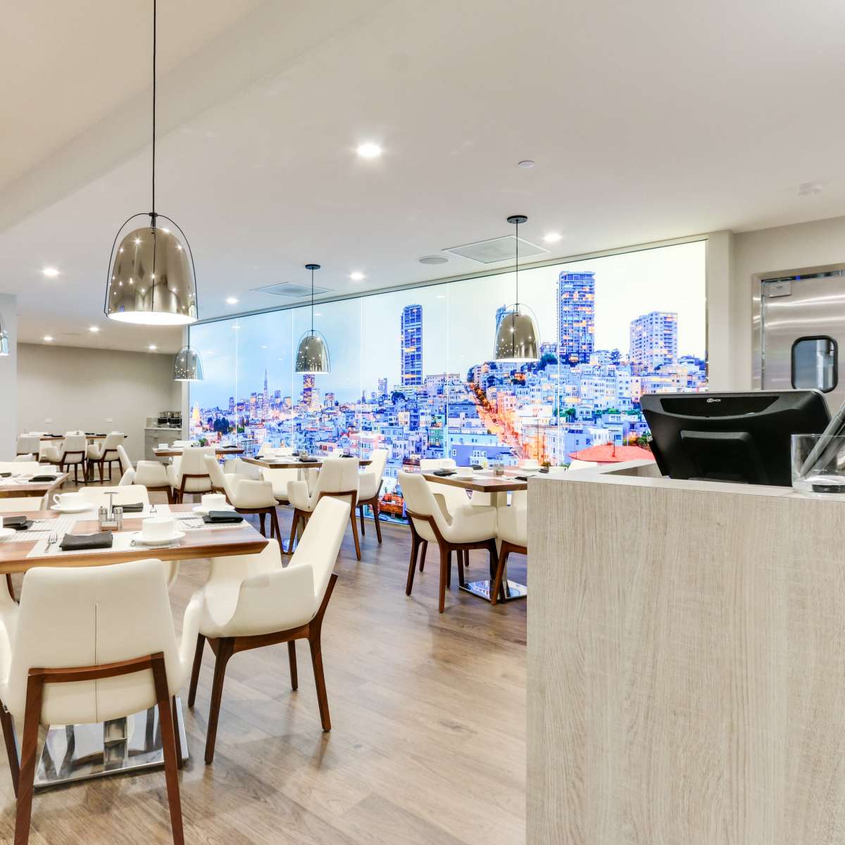A modern restaurant interior with wooden flooring, white tables, and chairs, illuminated by pendant lights, featuring a large digital cityscape mural on one wall.