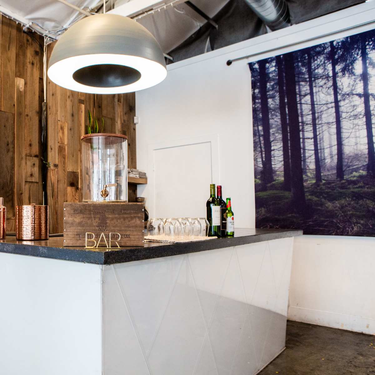 A modern bar counter made of wood and concrete, featuring a large pendant light and a selection of bottles and glasses neatly arranged on the surface.