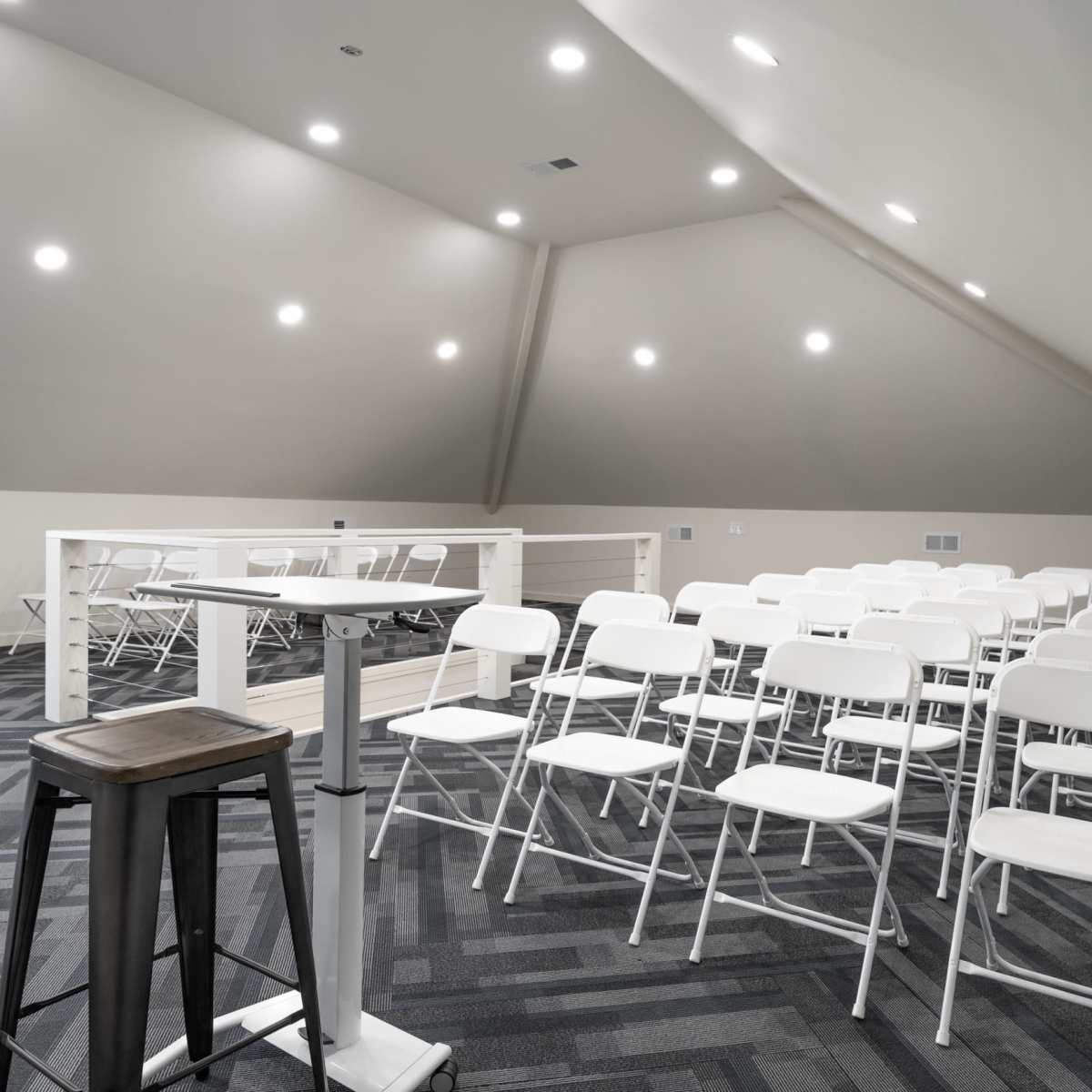 A spacious attic room features rows of white folding chairs set up for an event, with a small podium in the foreground.