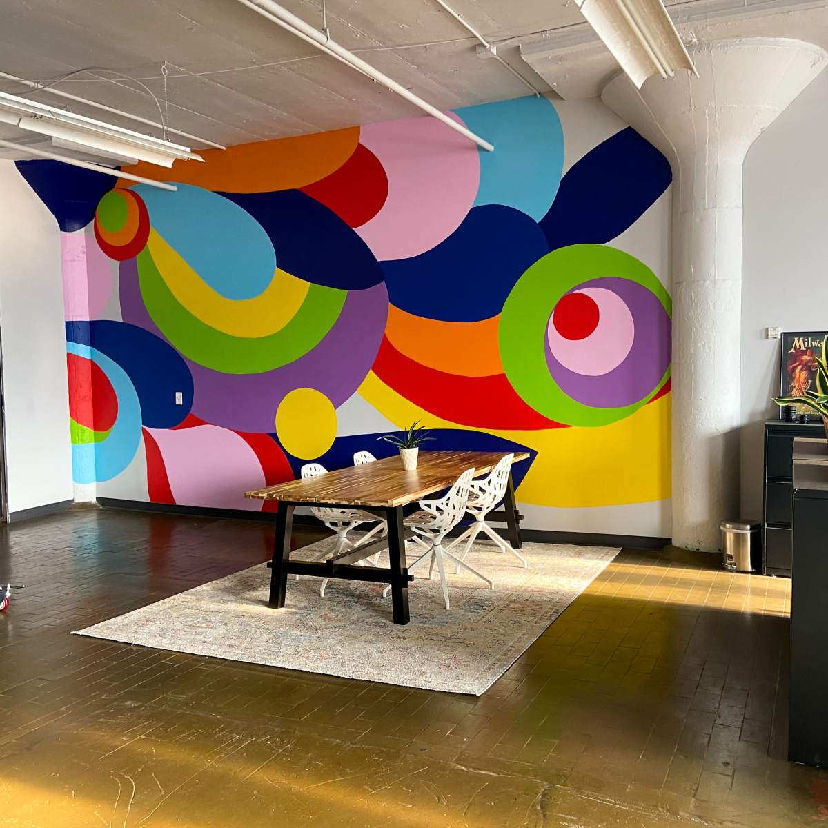 A brightly painted wall with abstract shapes in various colors, alongside a wooden table and chairs on a patterned rug in a spacious room with wooden flooring.