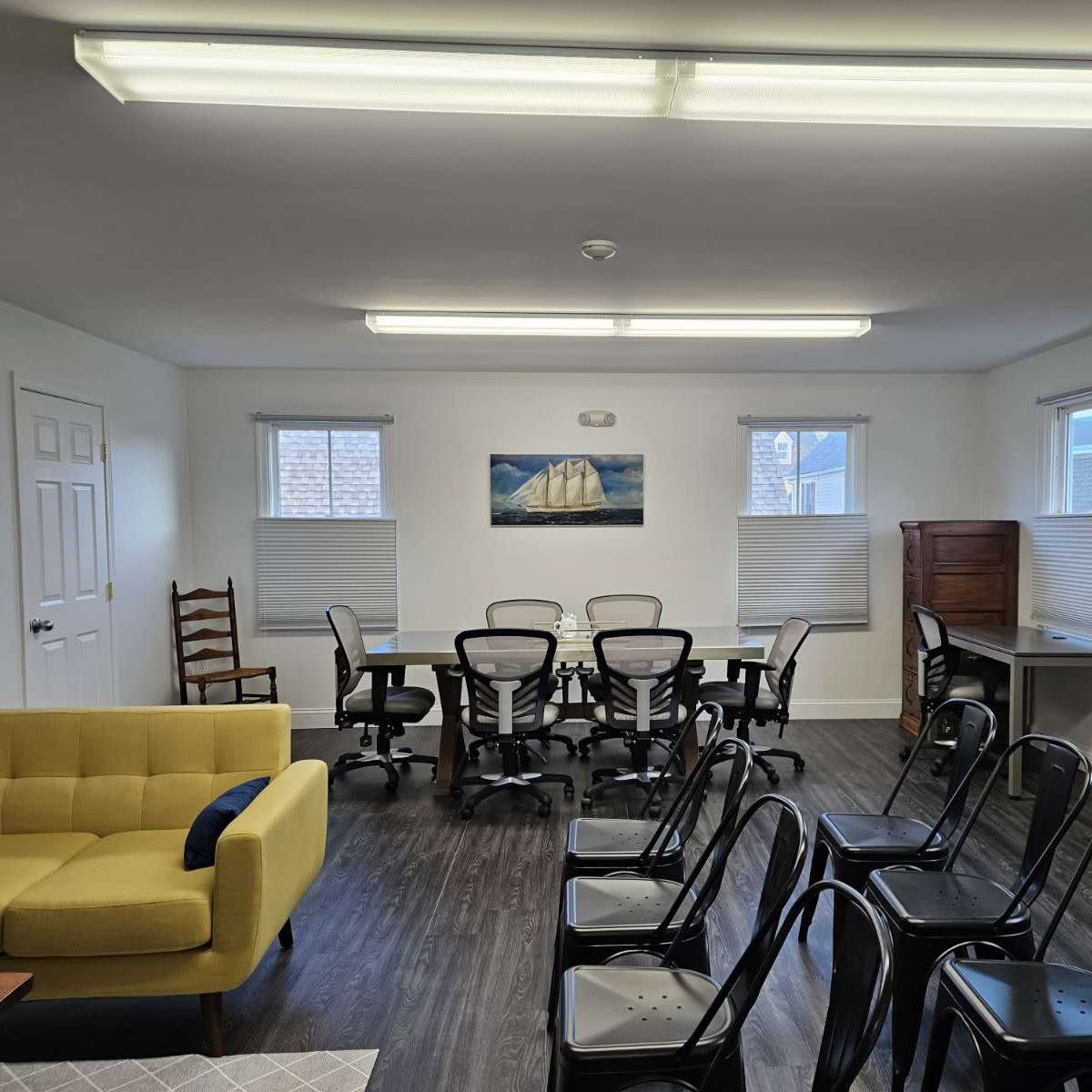 A well-lit meeting room with a large table surrounded by rolling chairs, a yellow sofa in the corner, and framed nautical artwork on the walls.