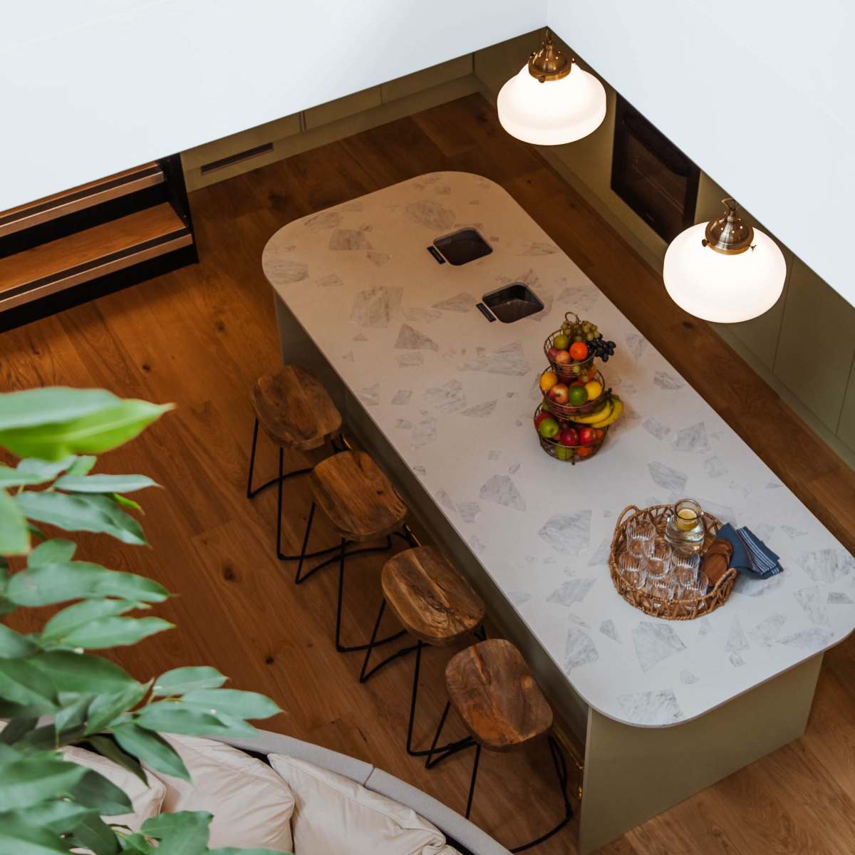 An overhead view of a modern kitchen featuring a large, oval marble dining table with wooden stools and decorative fruit arrangements, illuminated by two pendant lights.