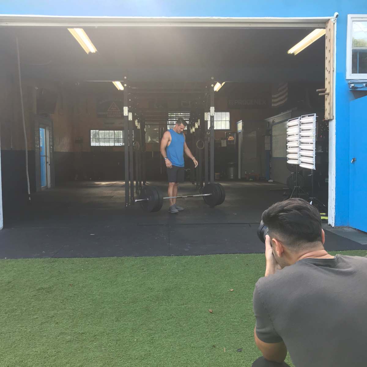 A man prepares to lift a barbell in a gym while another person photographs him from a distance.