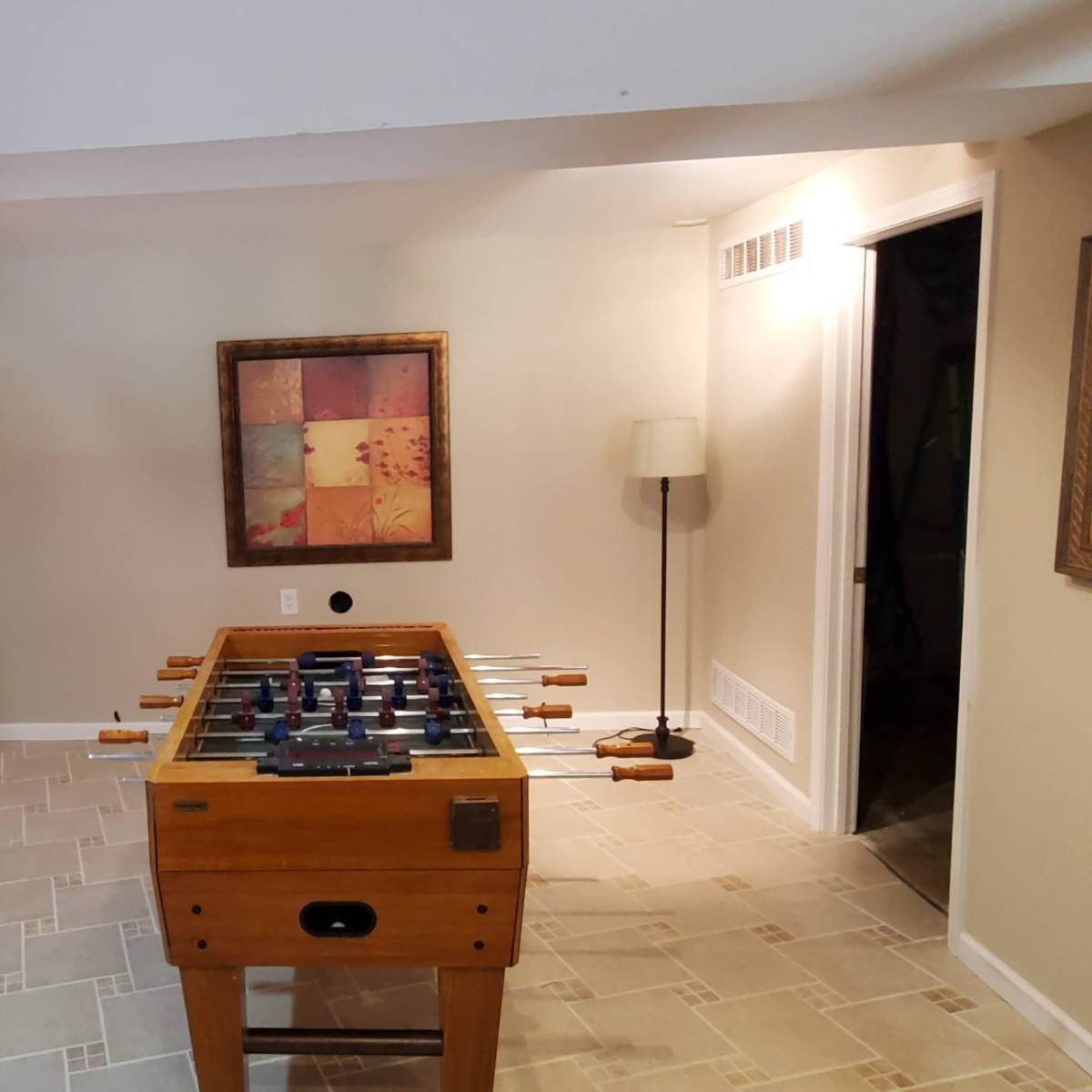 A room with a foosball table in the center, surrounded by shelves and a wall art piece, along with a lamp in the corner.