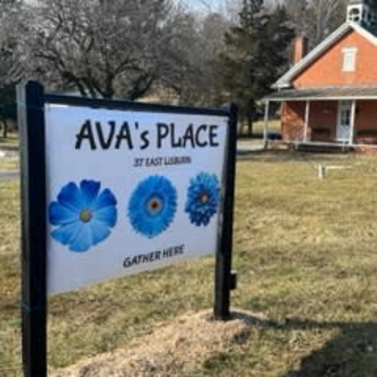 A sign for "Ava's Place," featuring blue flower graphics, in front of a brick building on a grassy area.