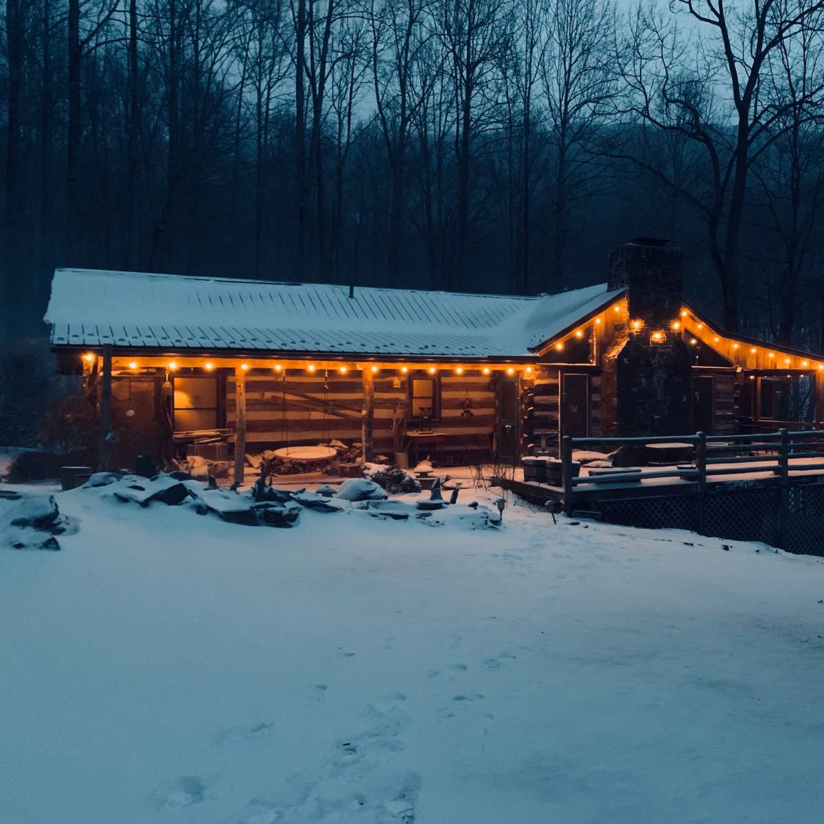 A cabin with string lights sits amidst a snowy landscape, surrounded by bare trees in the evening light.