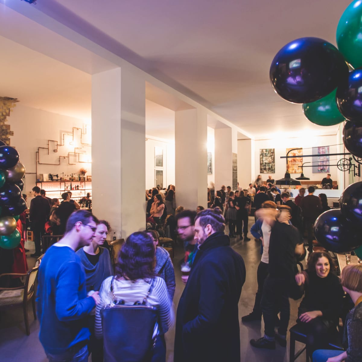 A crowd of people socializes at a lively indoor party, surrounded by decorations including large green and black balloons.