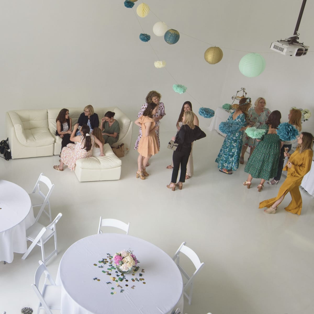 A bright, spacious event room features two round tables decorated with floral arrangements, while guests mingle and a cake display is set up in the background.