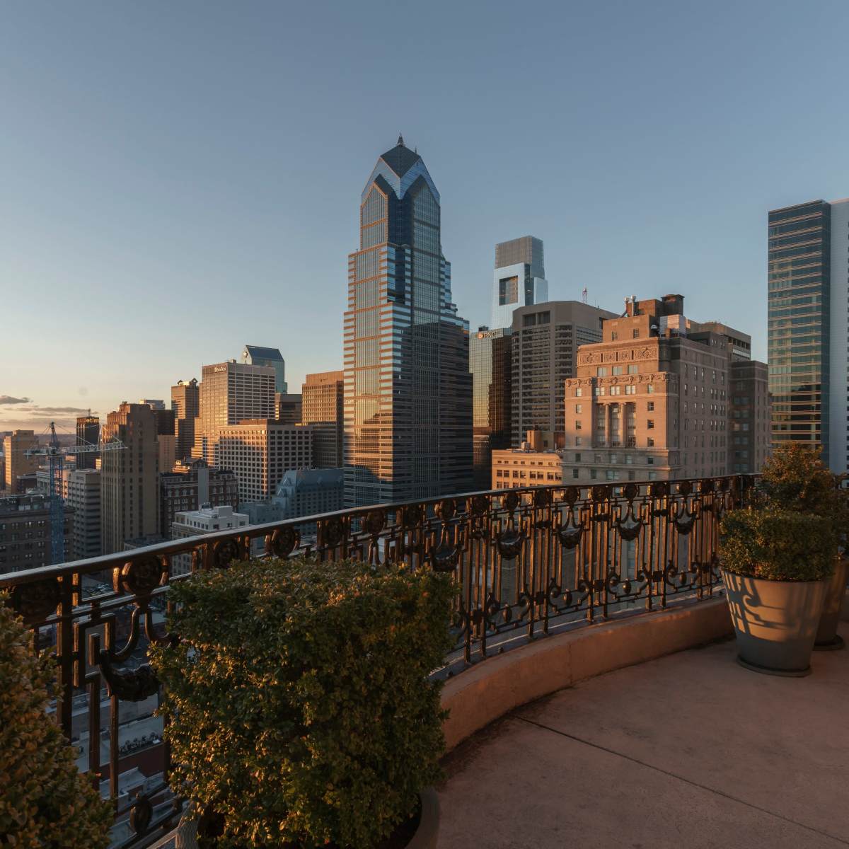 A balcony with potted plants overlooking a skyline of tall buildings in a city at sunset.