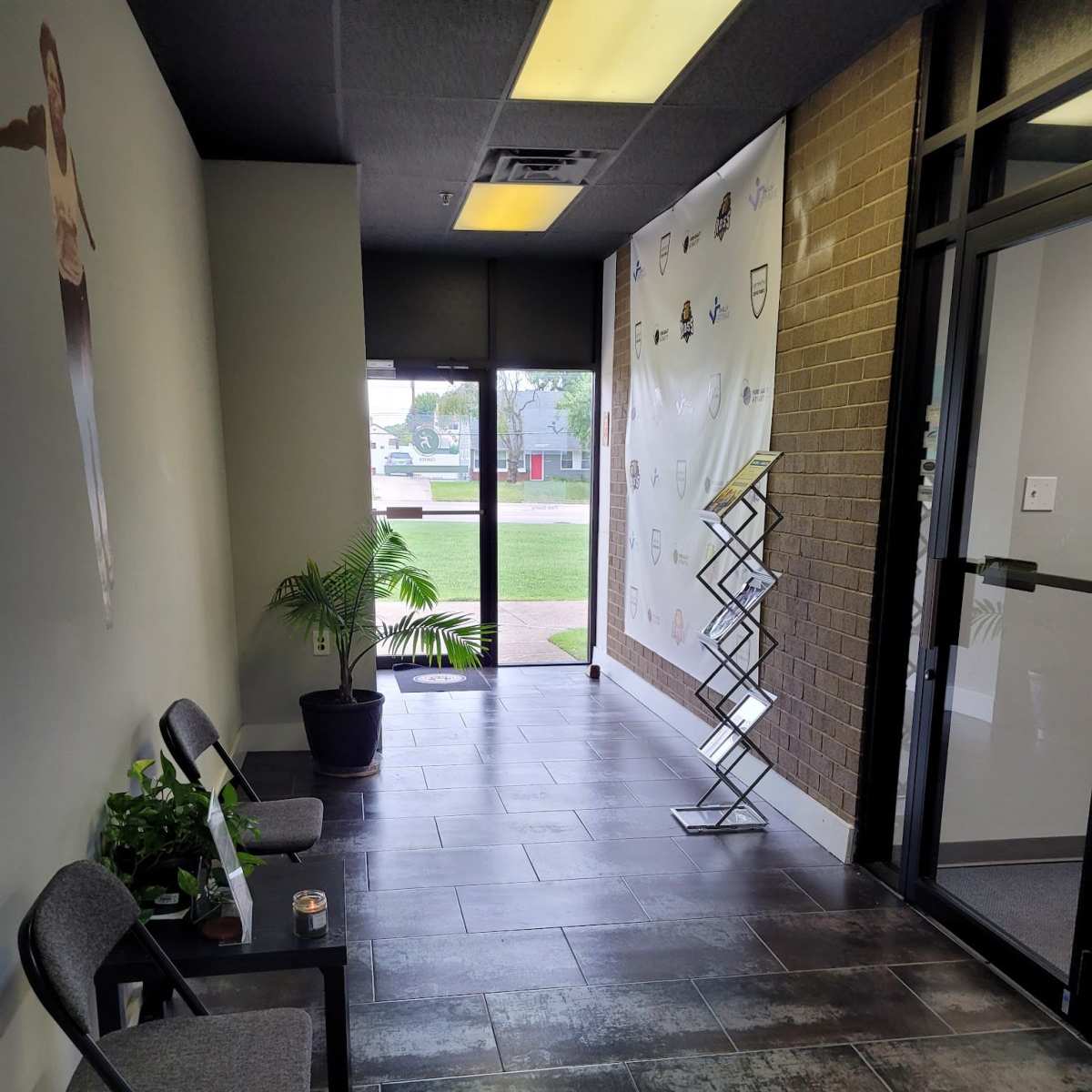 A narrow hallway with chairs, a potted plant, and a wall displaying logos, leading to a glass door with an exterior view.