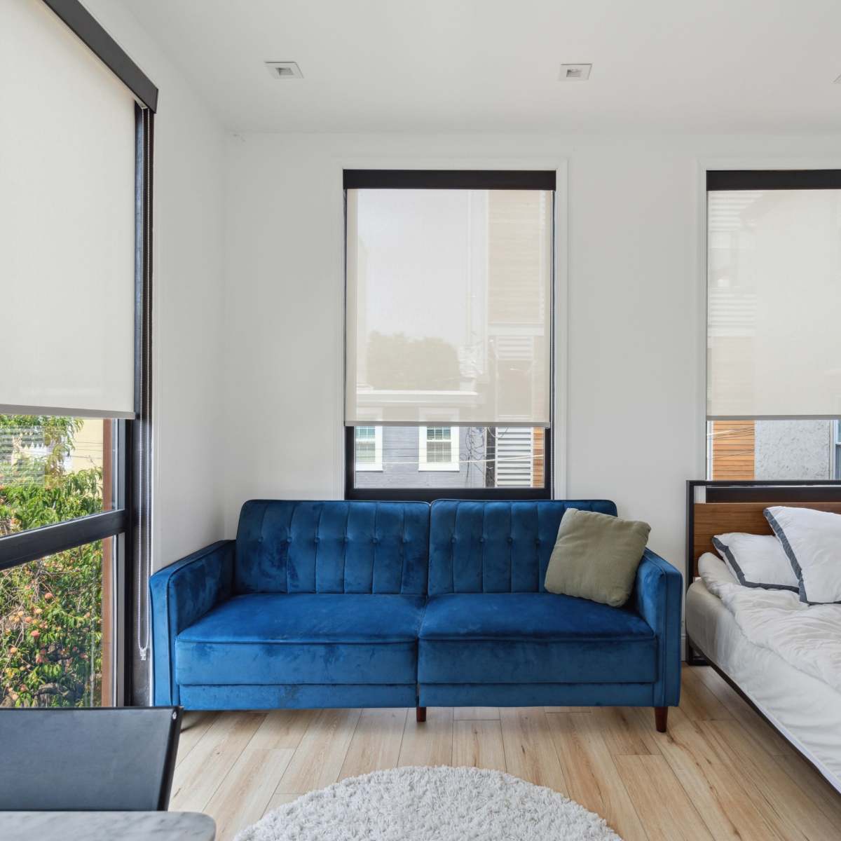 A bright, minimalist bedroom featuring a blue velvet sofa, a wooden bed with unmade white linens, and large windows with light curtains.