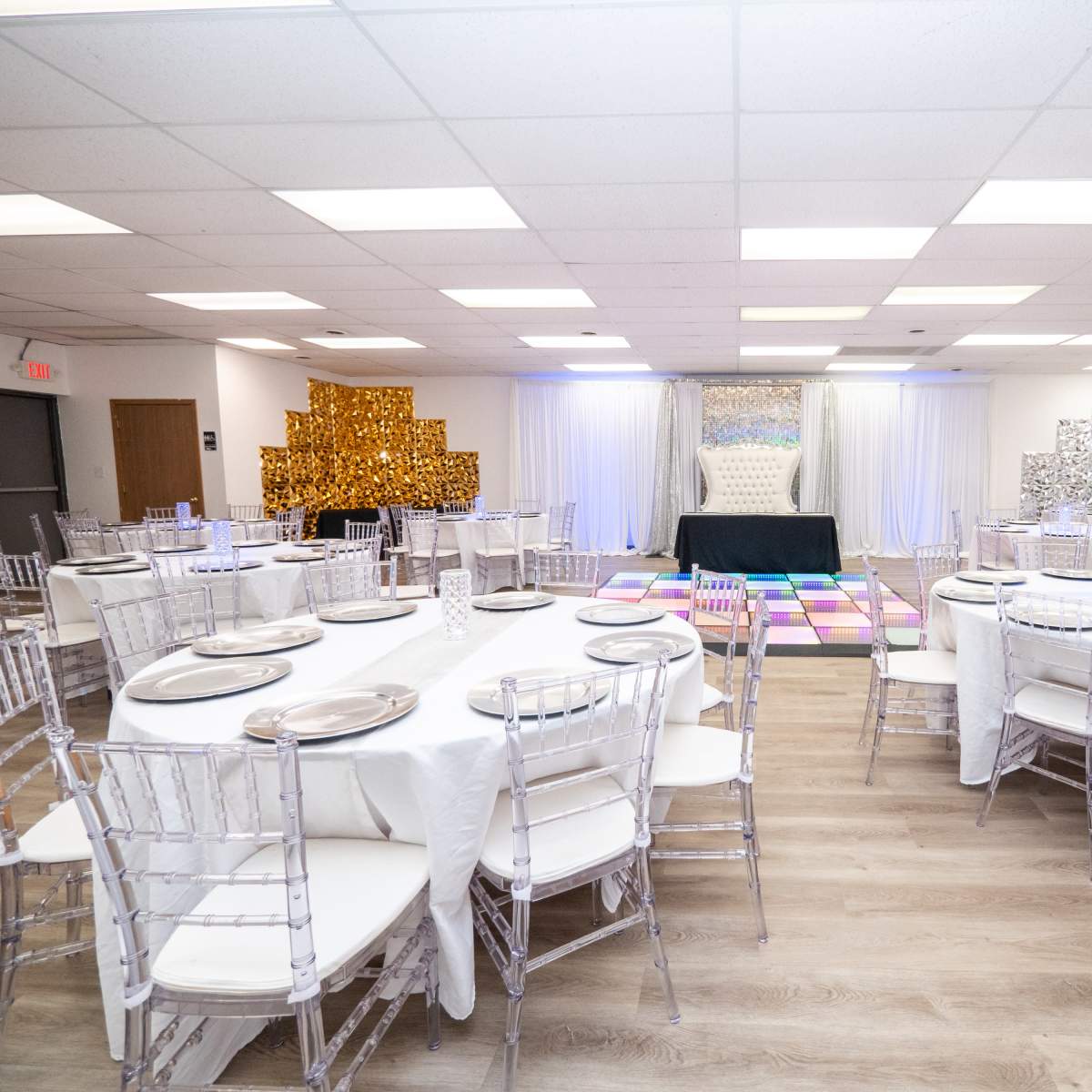 A banquet hall set up with round tables and clear chiavari chairs, featuring decorative backdrops on the walls and a central stage area.