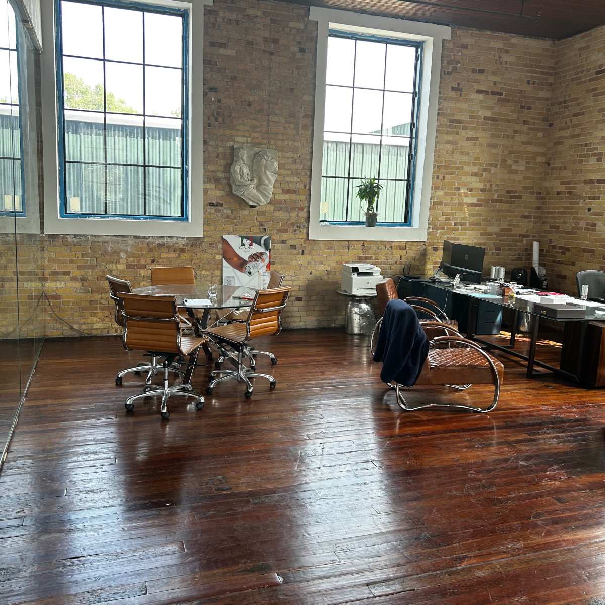 A spacious office with wooden floors, large windows, a meeting table with chairs, and a desk area in the corner.