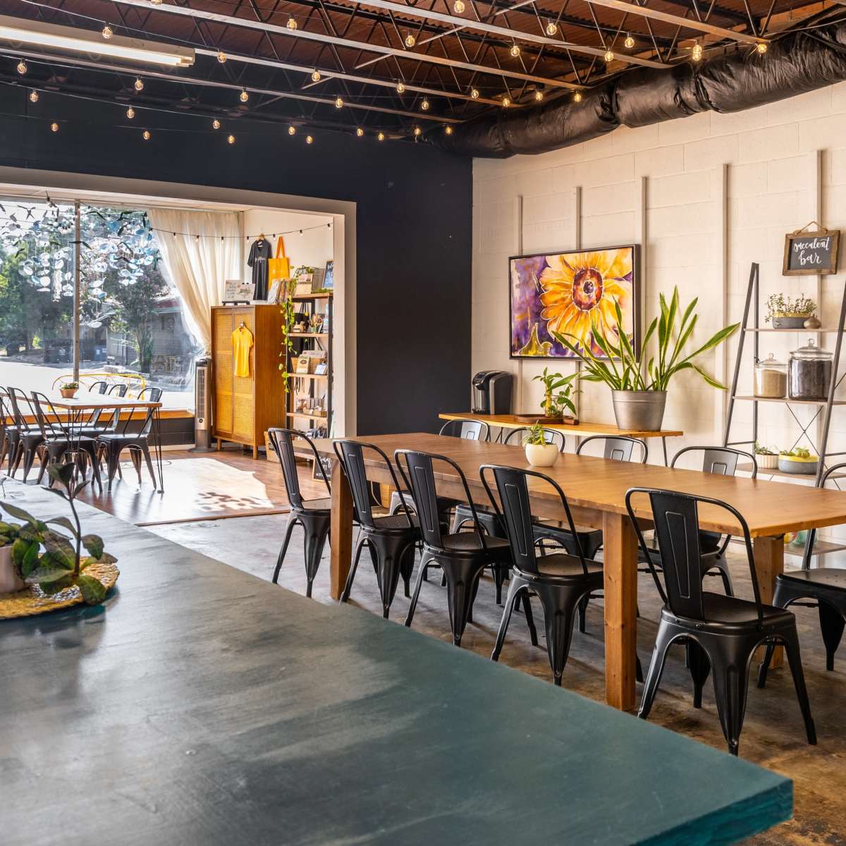 A bright, spacious interior of a café with wooden tables, industrial-style chairs, and large windows allowing natural light to enter.