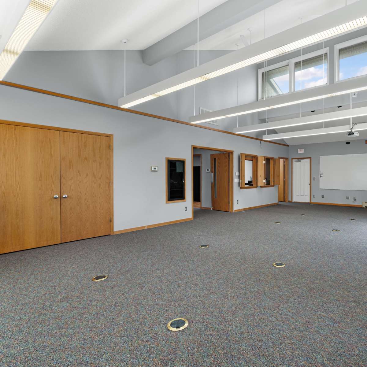 An empty room with gray walls and carpet, featuring large windows, ceiling lights, and several doors.