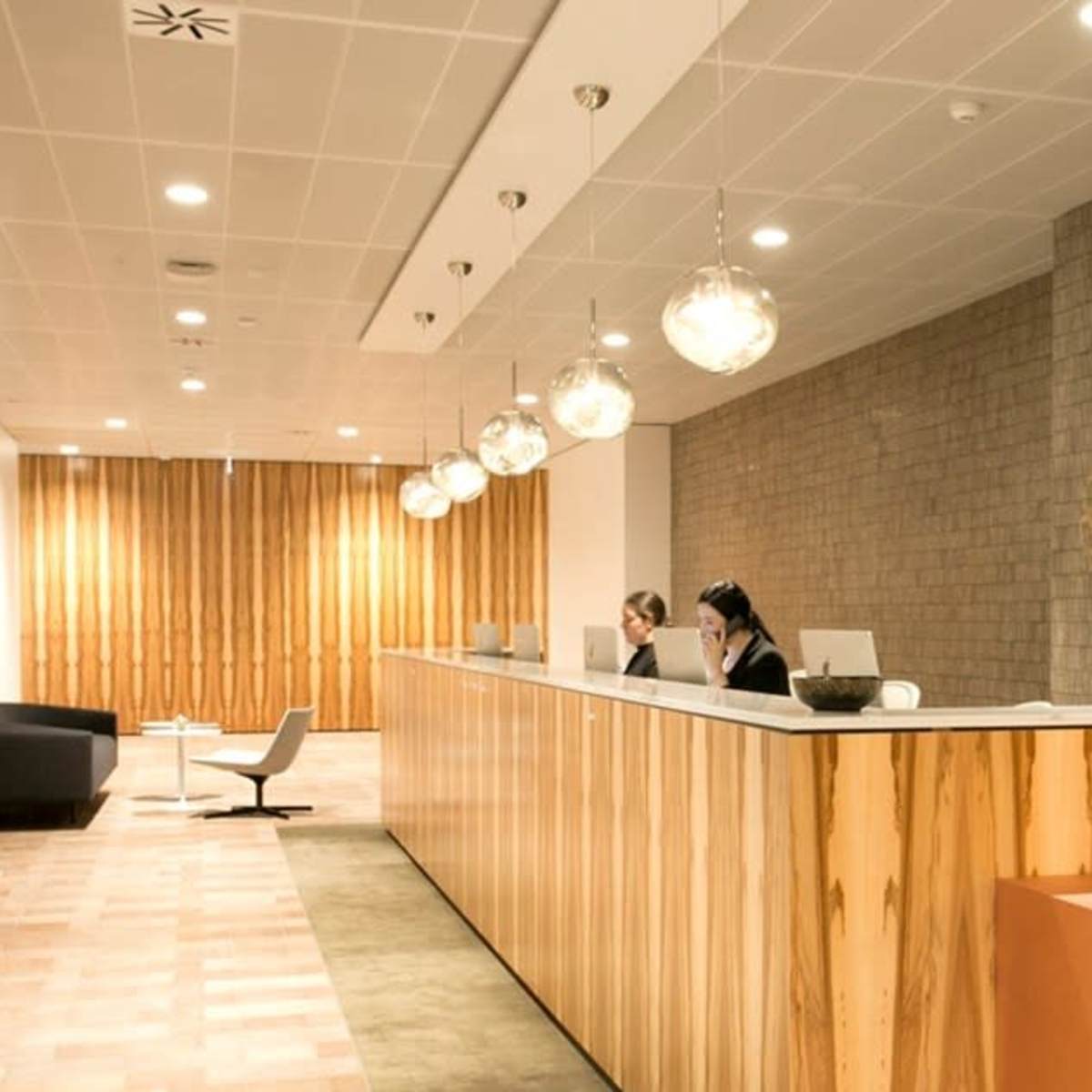 A modern reception area with a wooden desk, two staff members seated behind it, and pendant lighting overhead.