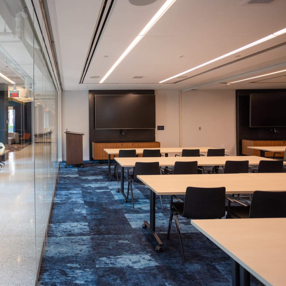 A modern conference room with glass walls, rows of tables and chairs, and large screens on the opposite wall.