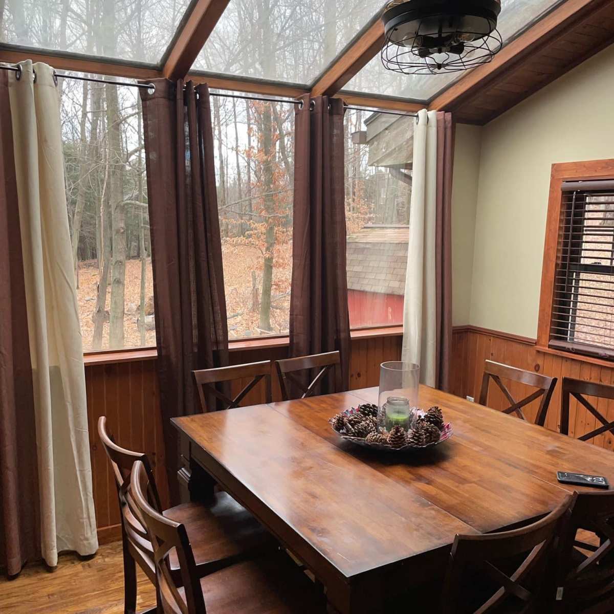 A wooden dining table surrounded by chairs is positioned in a sunlit room with large windows overlooking a wooded area.