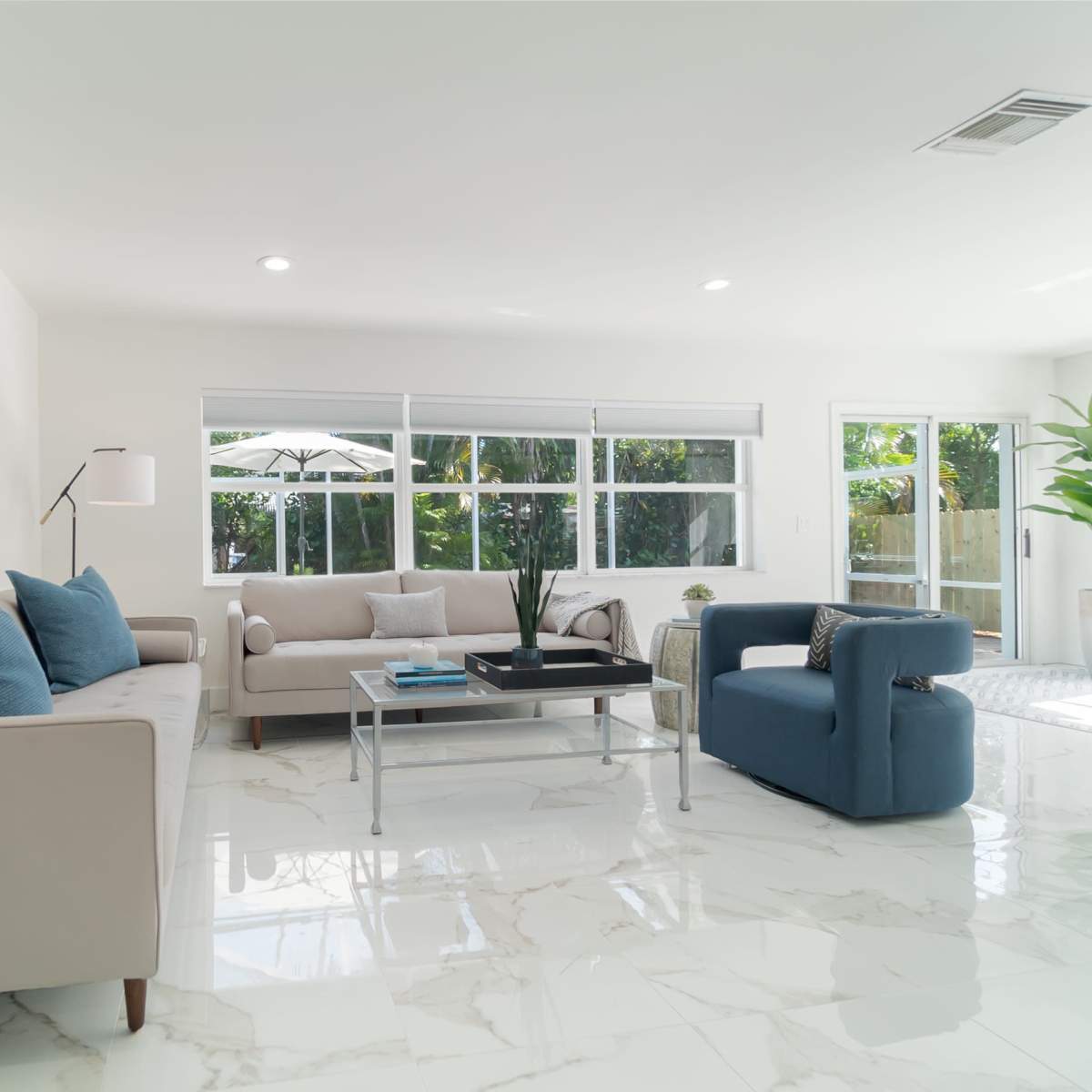 A modern living room features a light-colored sofa, a blue armchair, and large windows that overlook a patio with greenery.