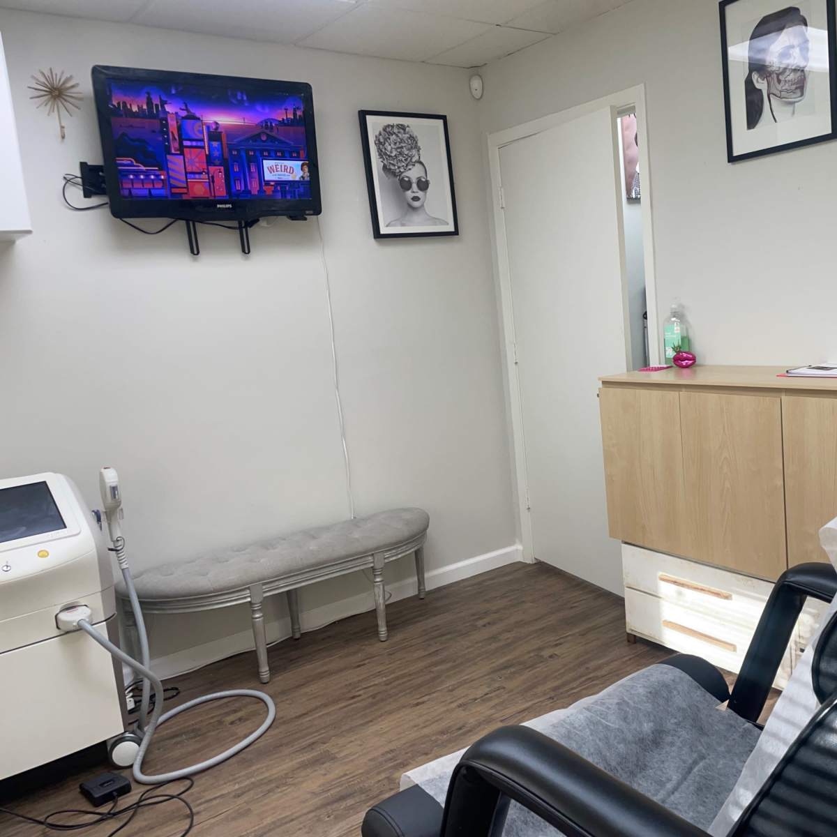 A small treatment room with a black reclining chair, a wooden cabinet, a television mounted on the wall, and a decorative wall featuring two framed pictures.