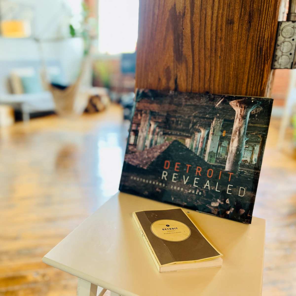 A coffee table holds a magazine titled "Detroit Revealed" next to a notebook in a room featuring hardwood floors and minimal furniture.
