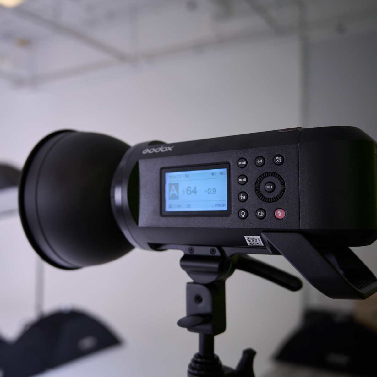 A studio strobe light is mounted on a stand, with its digital display showing settings, and softboxes are visible in the background.