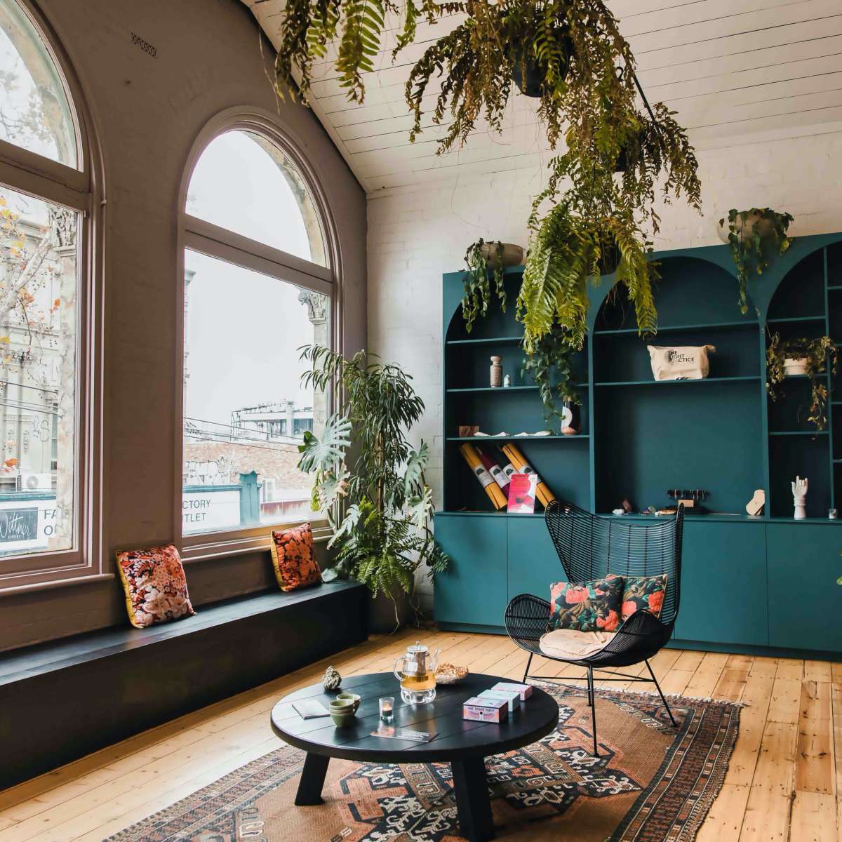 The room features large arched windows, a dark green shelving unit, a circular coffee table, and various potted plants.