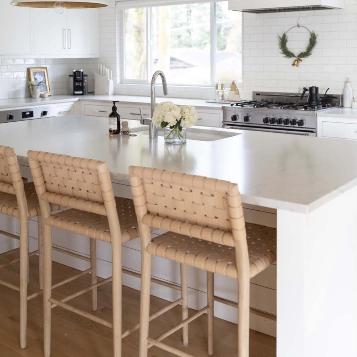 A modern kitchen features a large island with wicker bar stools, pendant lights, and a window overlooking an outdoor view.