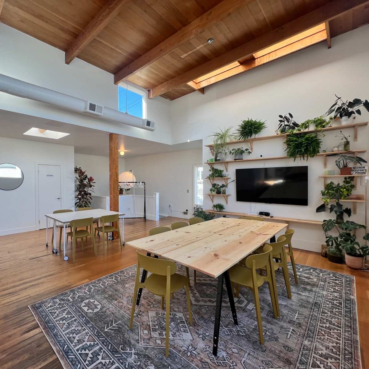 A spacious room with a dining table surrounded by green chairs, a wall-mounted television, and plants on shelves, with a kitchen area visible in the background.