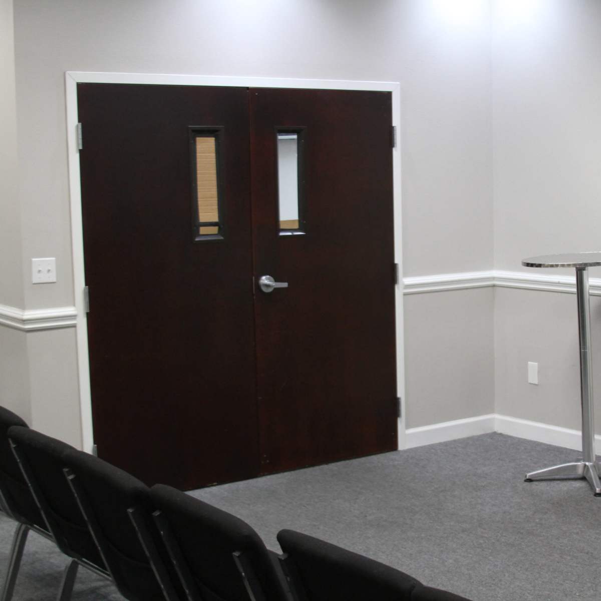 A small meeting room featuring a dark wooden door, a podium, a standing fan, and a television mounted on the wall, with rows of chairs arranged in front.