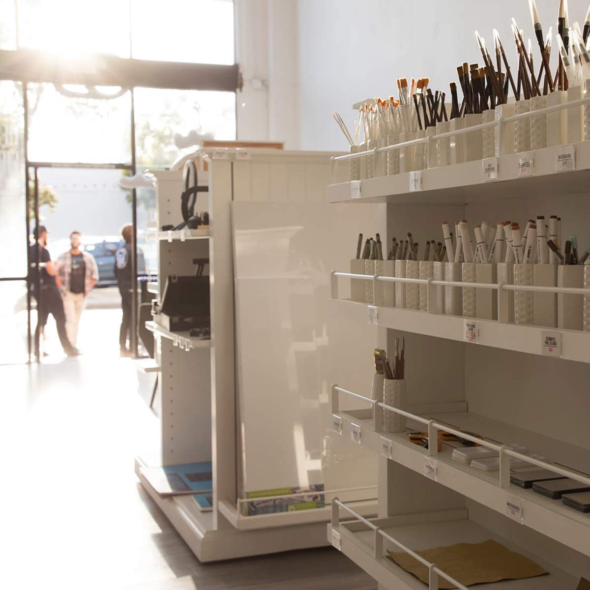 A well-organized store display with various pens and stationery items arranged on shelves, illuminated by natural light coming through the large front windows.