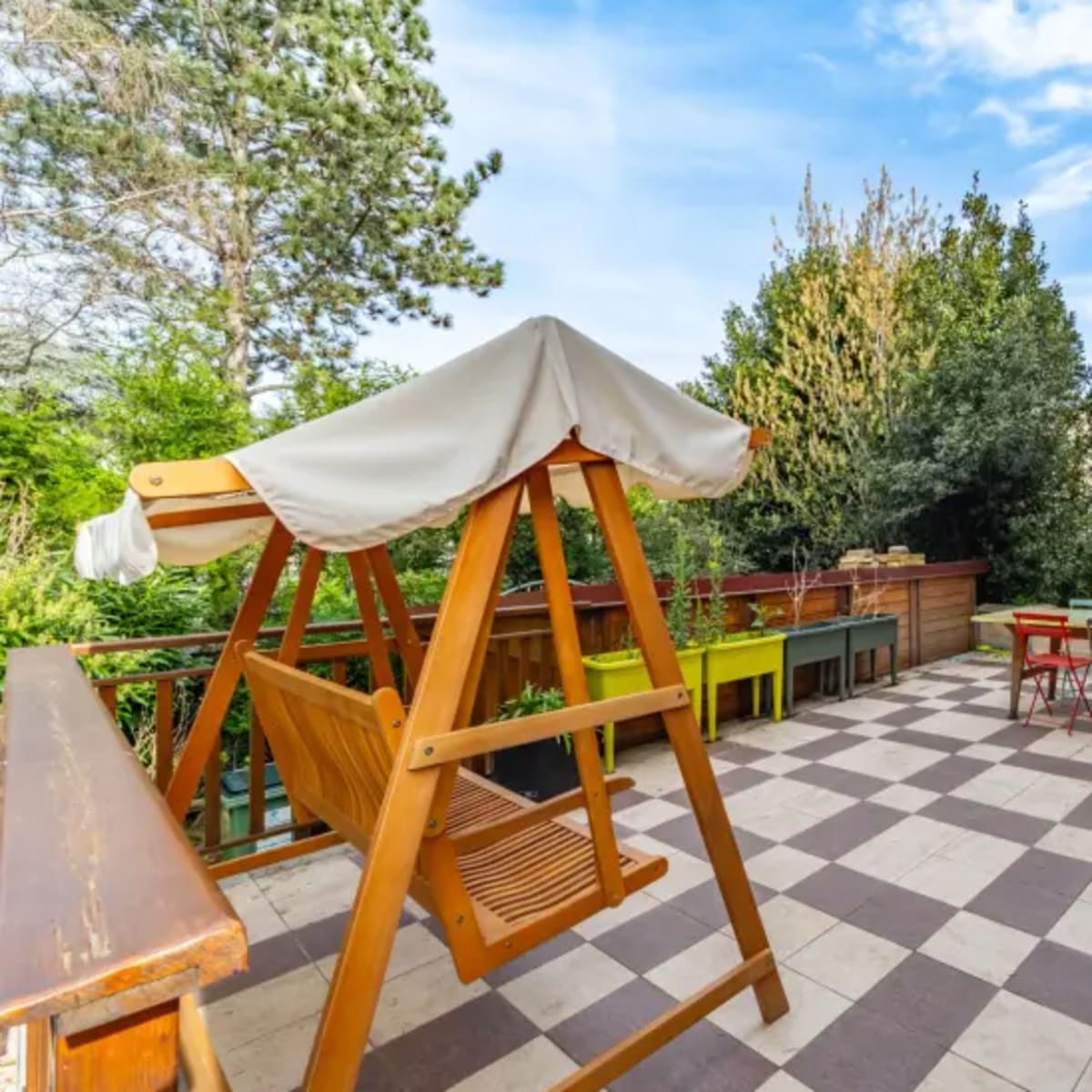 A patio area featuring a wooden swing, a table with chairs, and greenery surrounding the space.