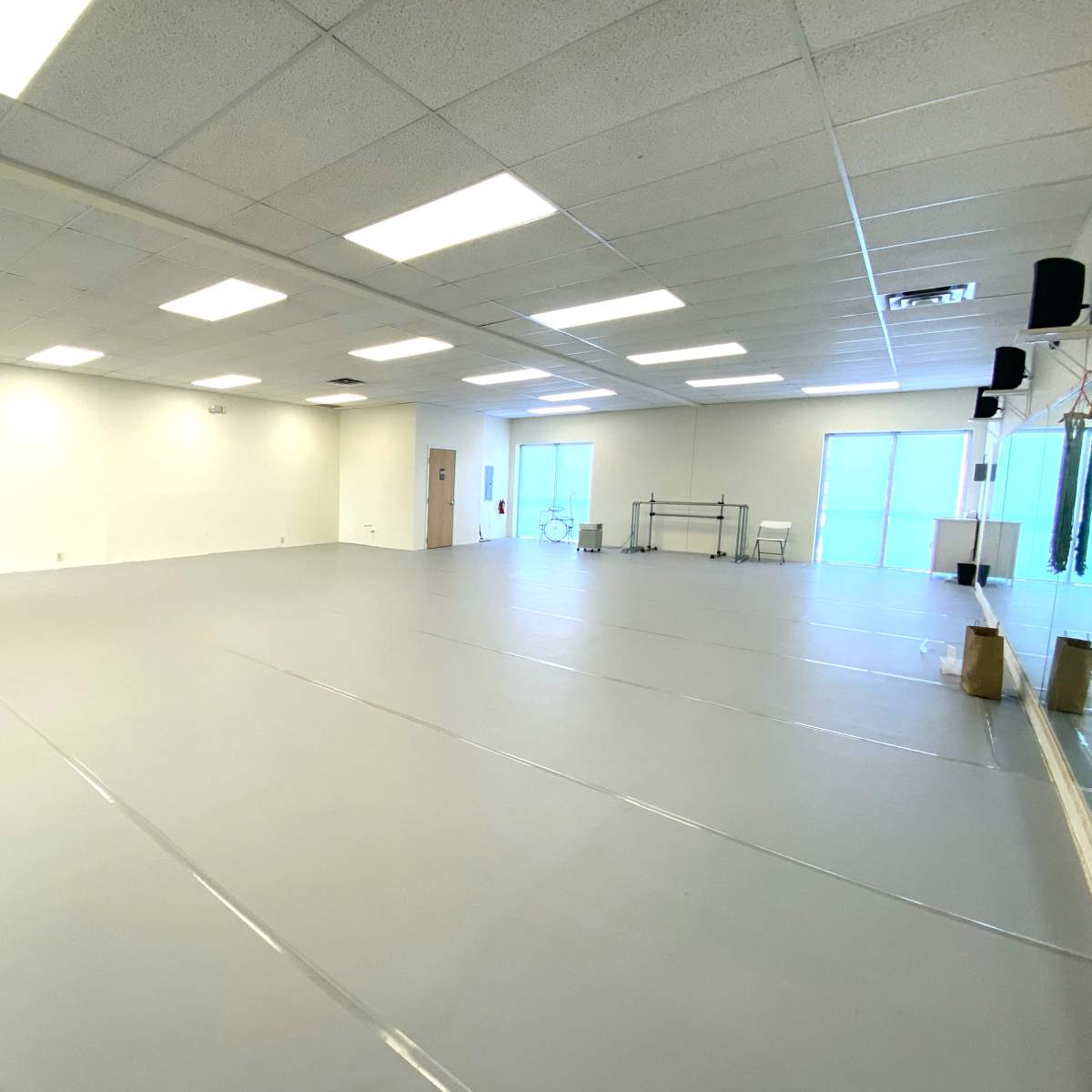 A large, empty studio space with grey flooring and white walls, featuring mirrors and bright overhead lighting.