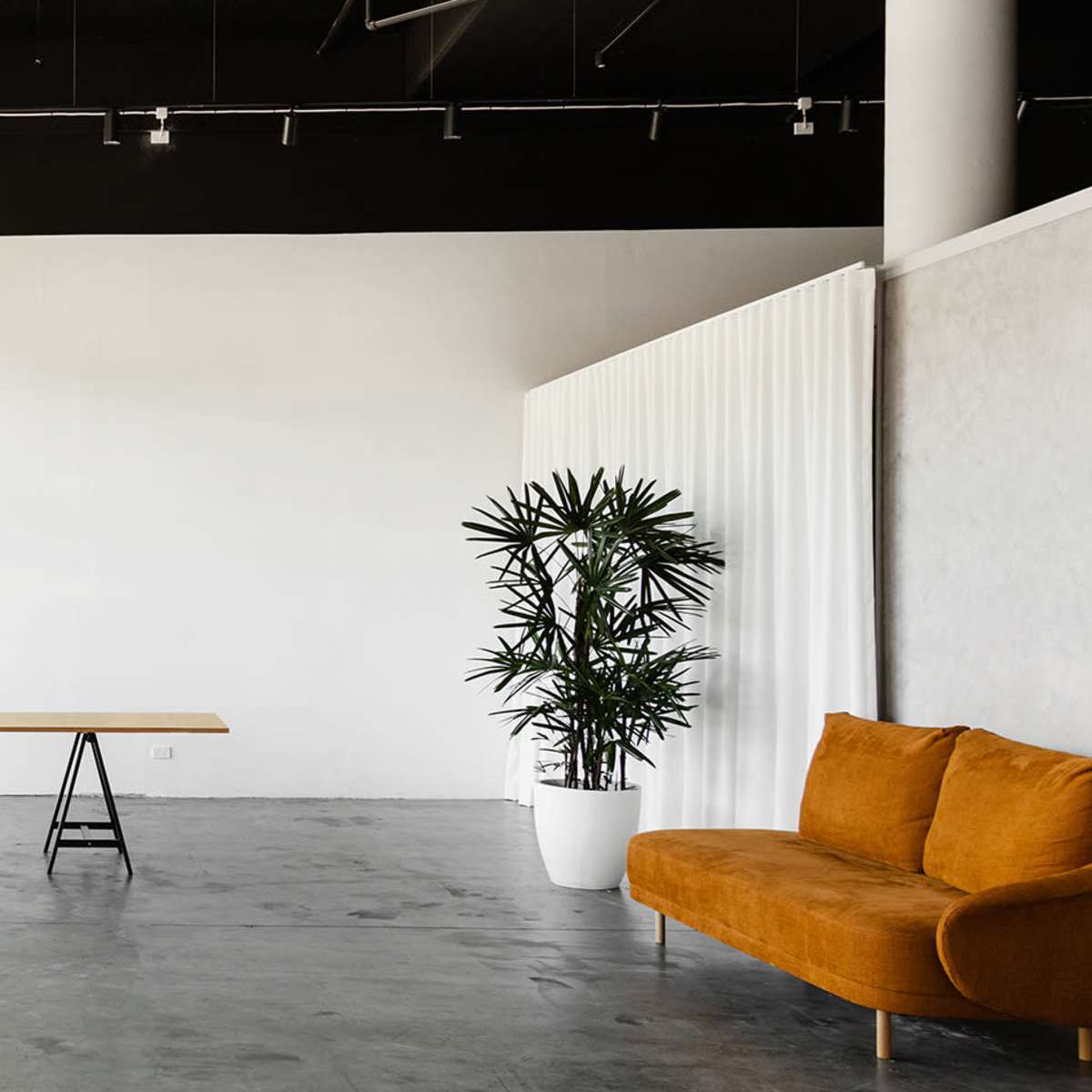 A spacious interior features a wooden table in the center, flanked by a gray sofa and an orange couch, with large windows and a plant in the corner.