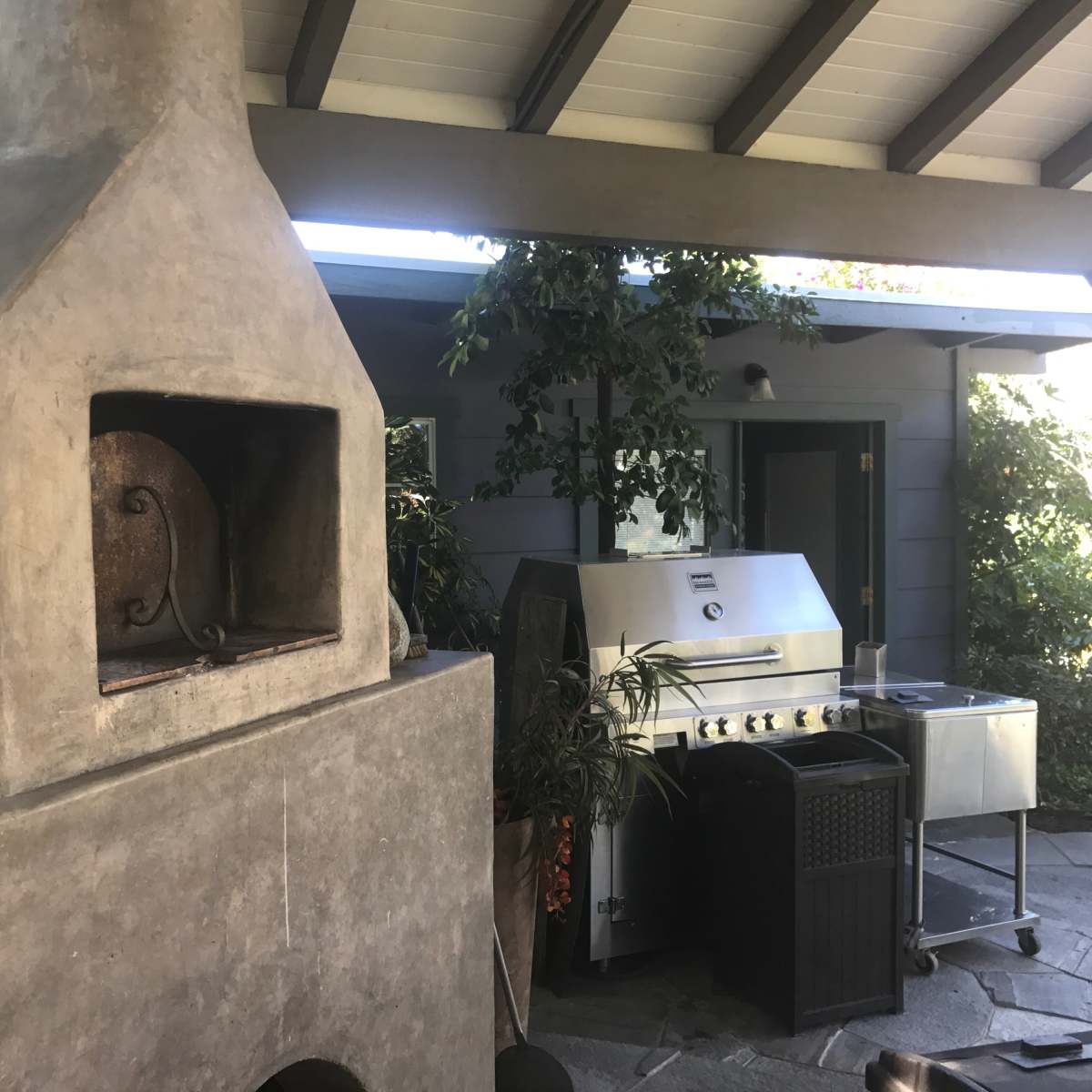 An outdoor kitchen area featuring a stone pizza oven and a stainless steel gas grill under a covered patio.