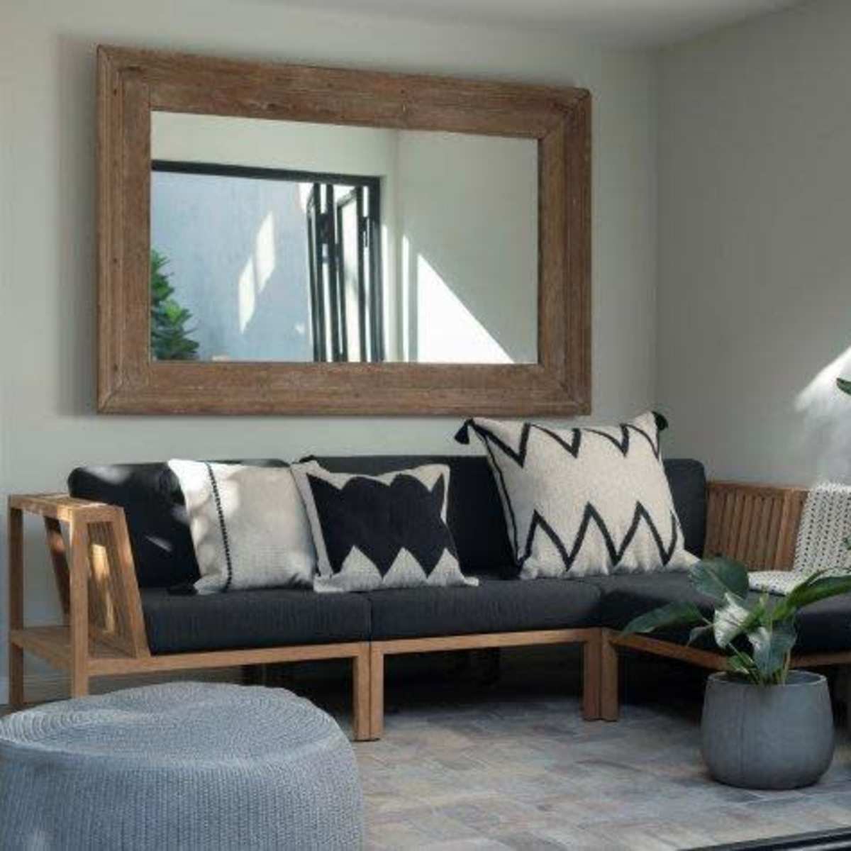 A minimalistic living space features a wooden-framed mirror, a sofa with patterned cushions, and potted plants arranged against a light-colored wall.