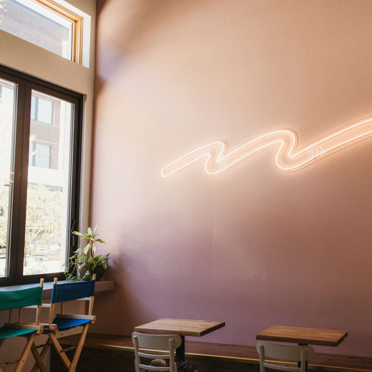 A minimalist café interior features a pink wall with a wavy neon light and wooden tables.