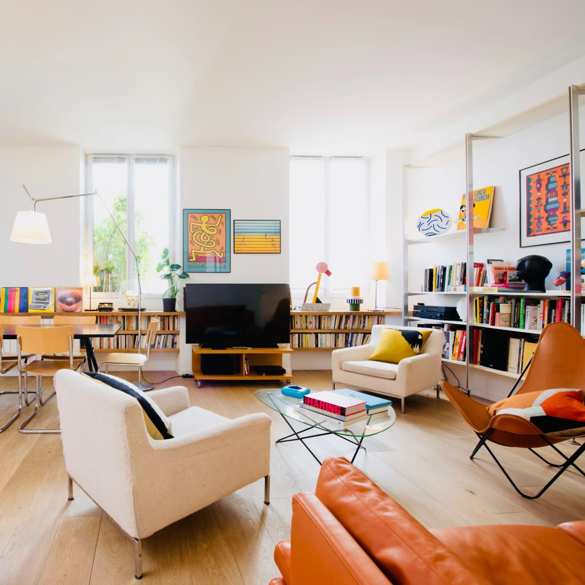 A modern living room with wooden flooring, a television, bookshelves filled with books, and various seating arrangements, including a white armchair and an orange leather chair.