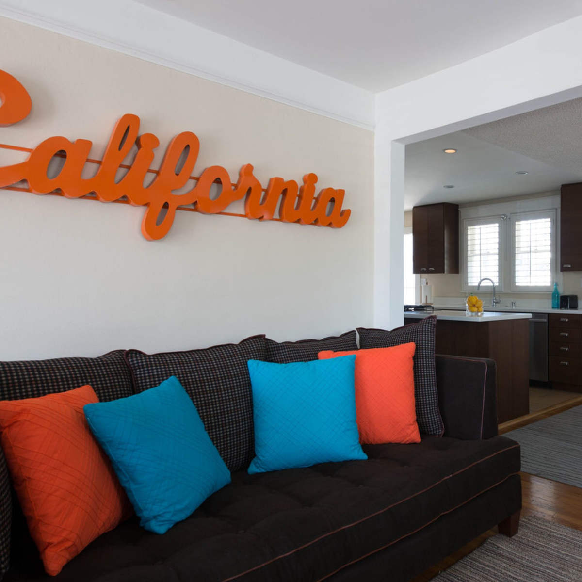 A living room features a dark couch with colorful pillows, a large orange "California" sign on the wall, and an adjacent dining area with a round table and white chairs.