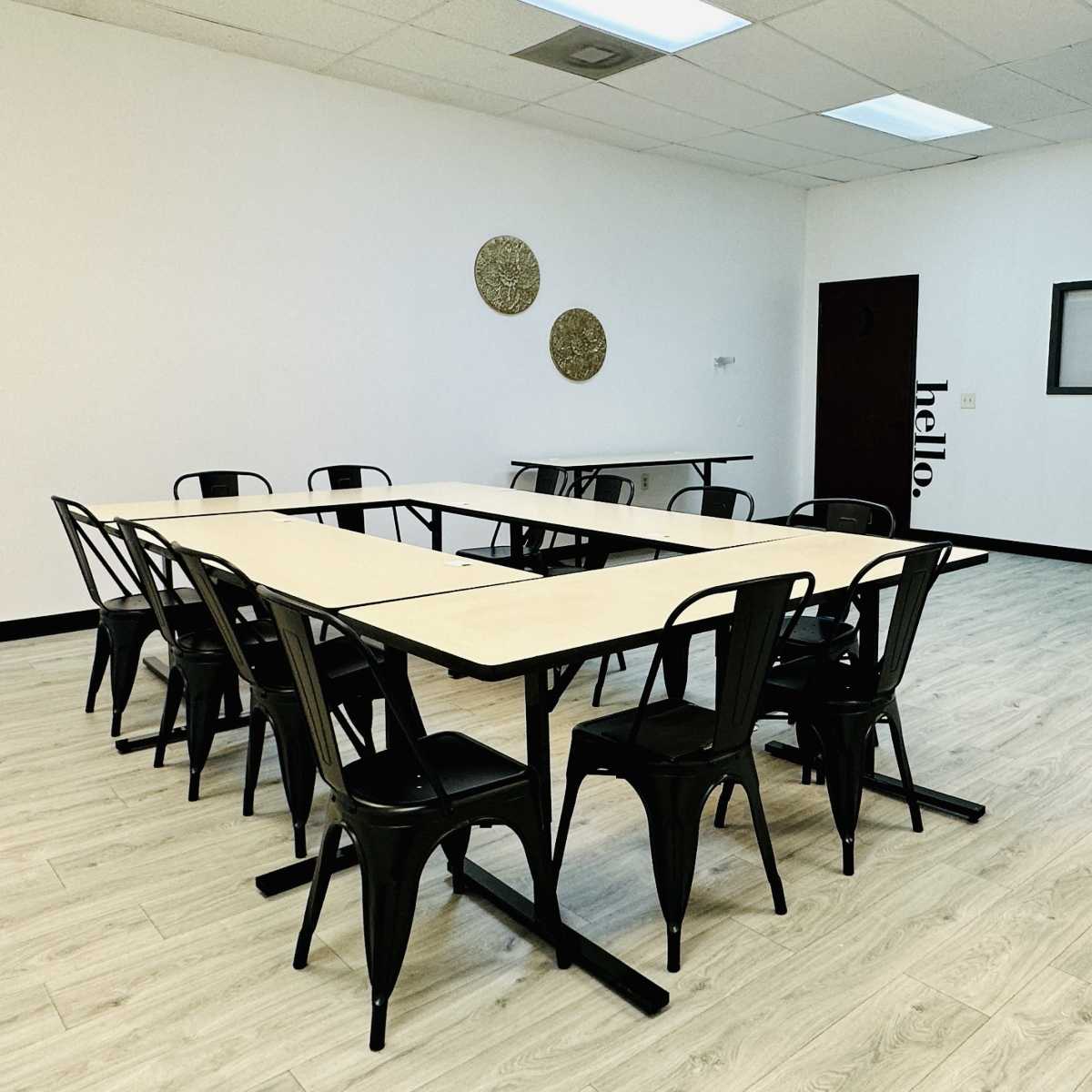 A meeting room features several tables arranged in a U-shape with black chairs and minimal decor.