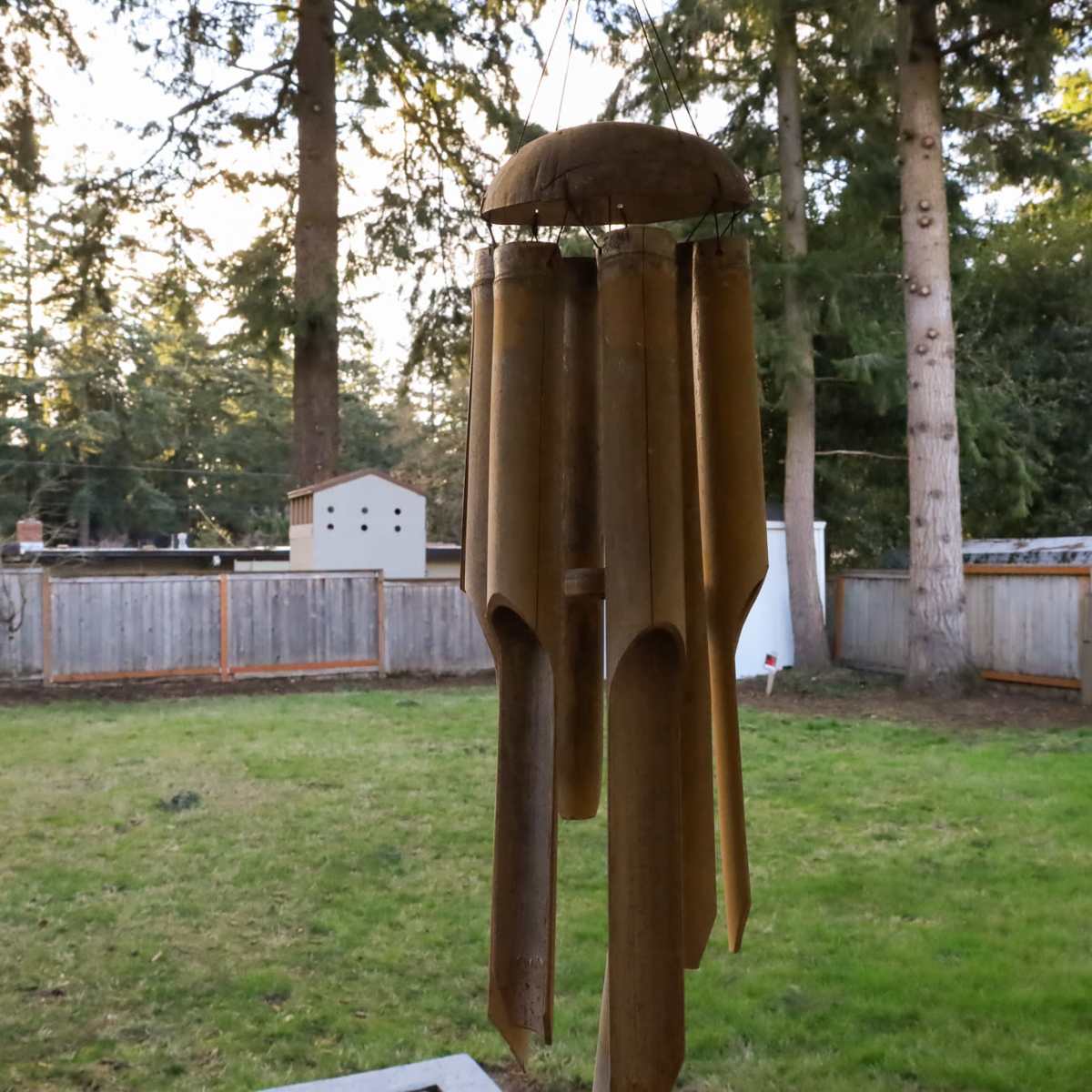 A wind chime made of wooden tubes hangs in a backyard surrounded by tall trees and a wooden fence.