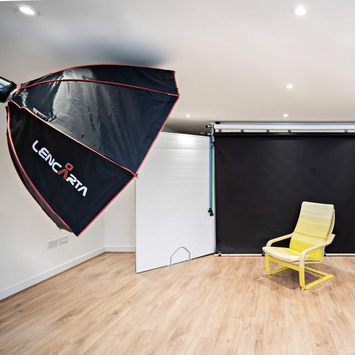 A yellow chair is positioned under a large softbox light in a minimalist photographic studio with black and white backdrop panels.