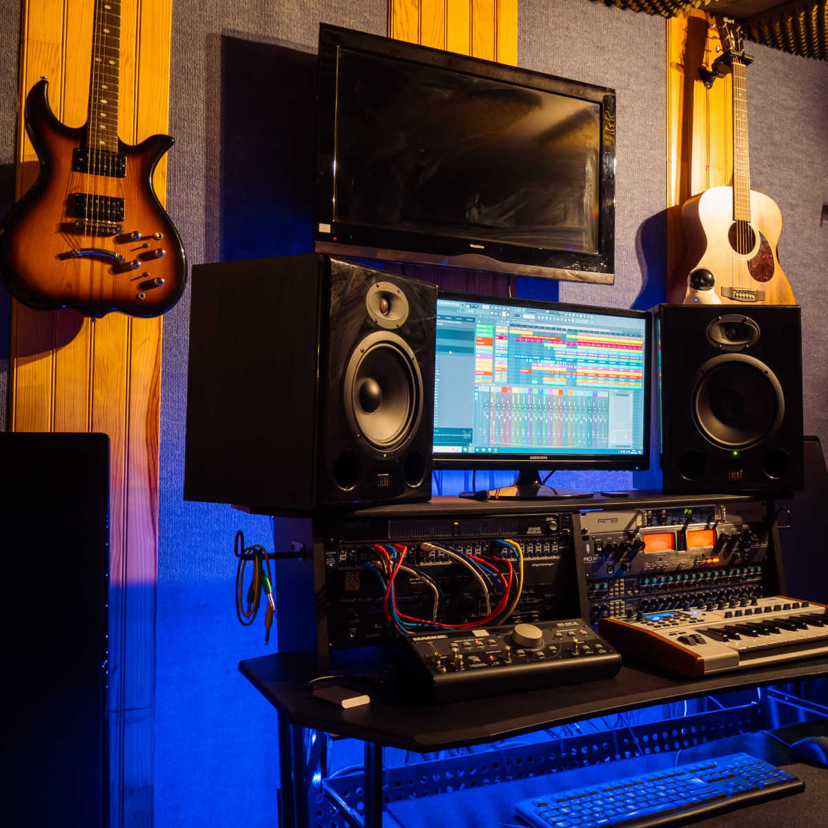 A music studio setup features a computer monitor displaying a digital audio workstation, flanked by speakers, a keyboard, and audio equipment, with guitars hanging on the wall.