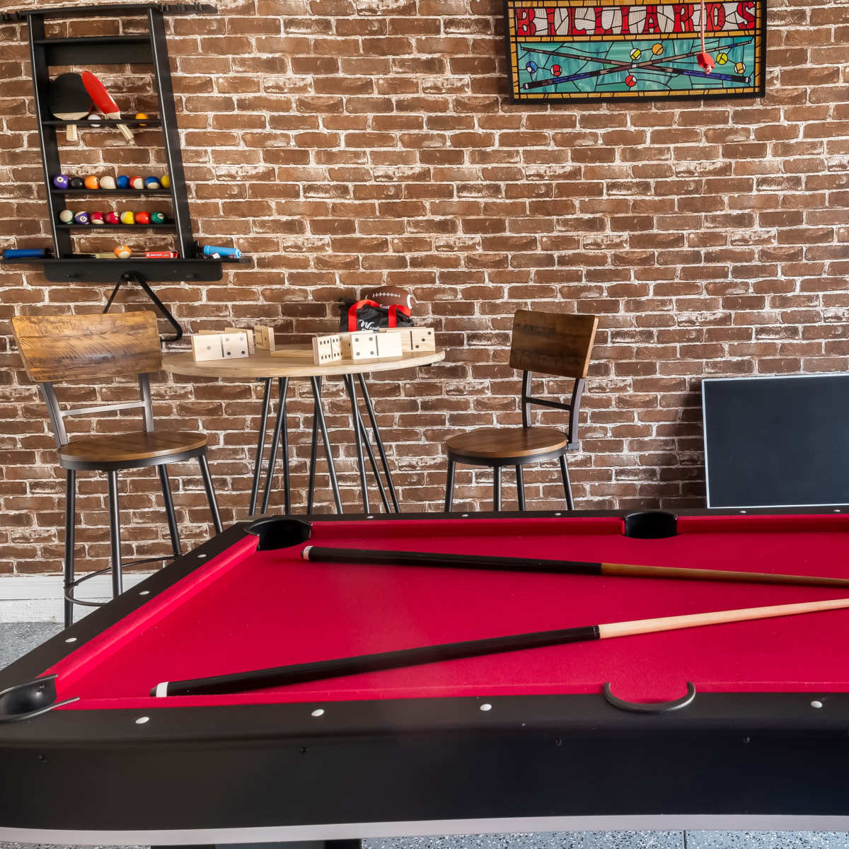 A red-felt billiard table is centered in a room with a brick wall, featuring a small table and chairs in the background.