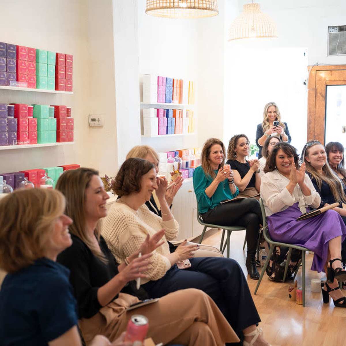 A group of fifteen women is seated and clapping in a brightly lit room with shelves displaying colorful products.