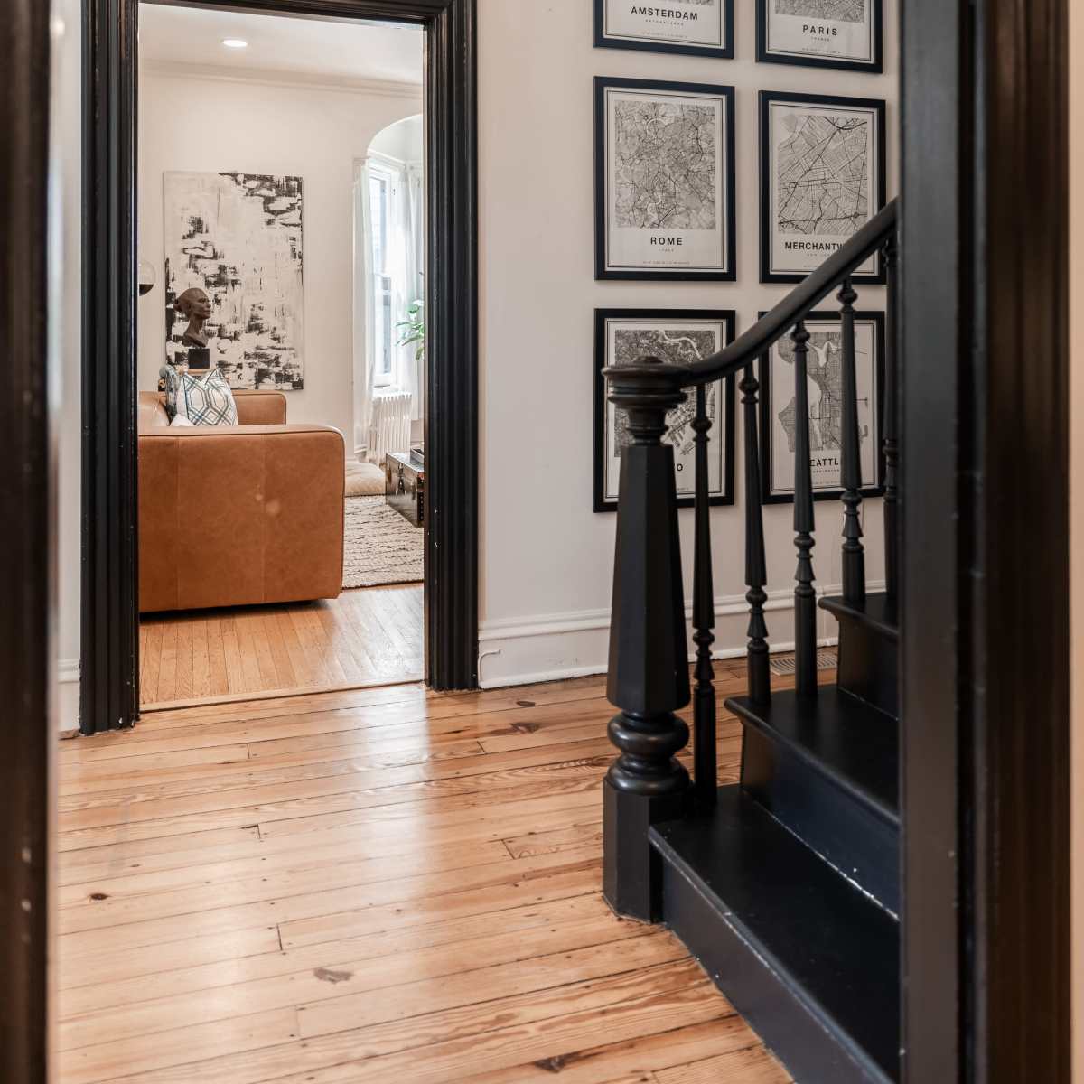 A hallway leads to a staircase, with framed maps on the wall and a view into a cozy living room.