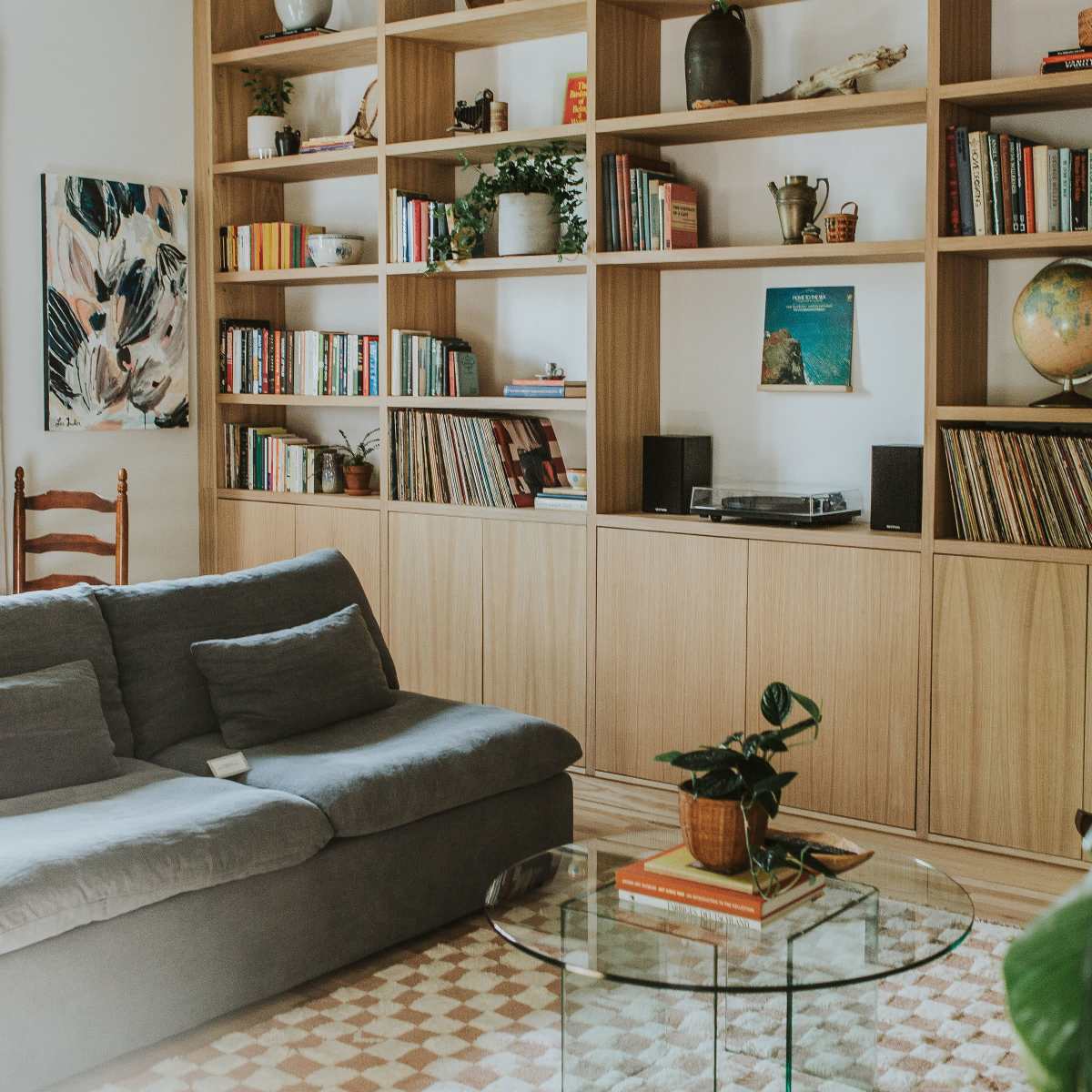 A cozy living room features a gray sofa, a glass coffee table, and wooden shelves filled with books and decorative items.