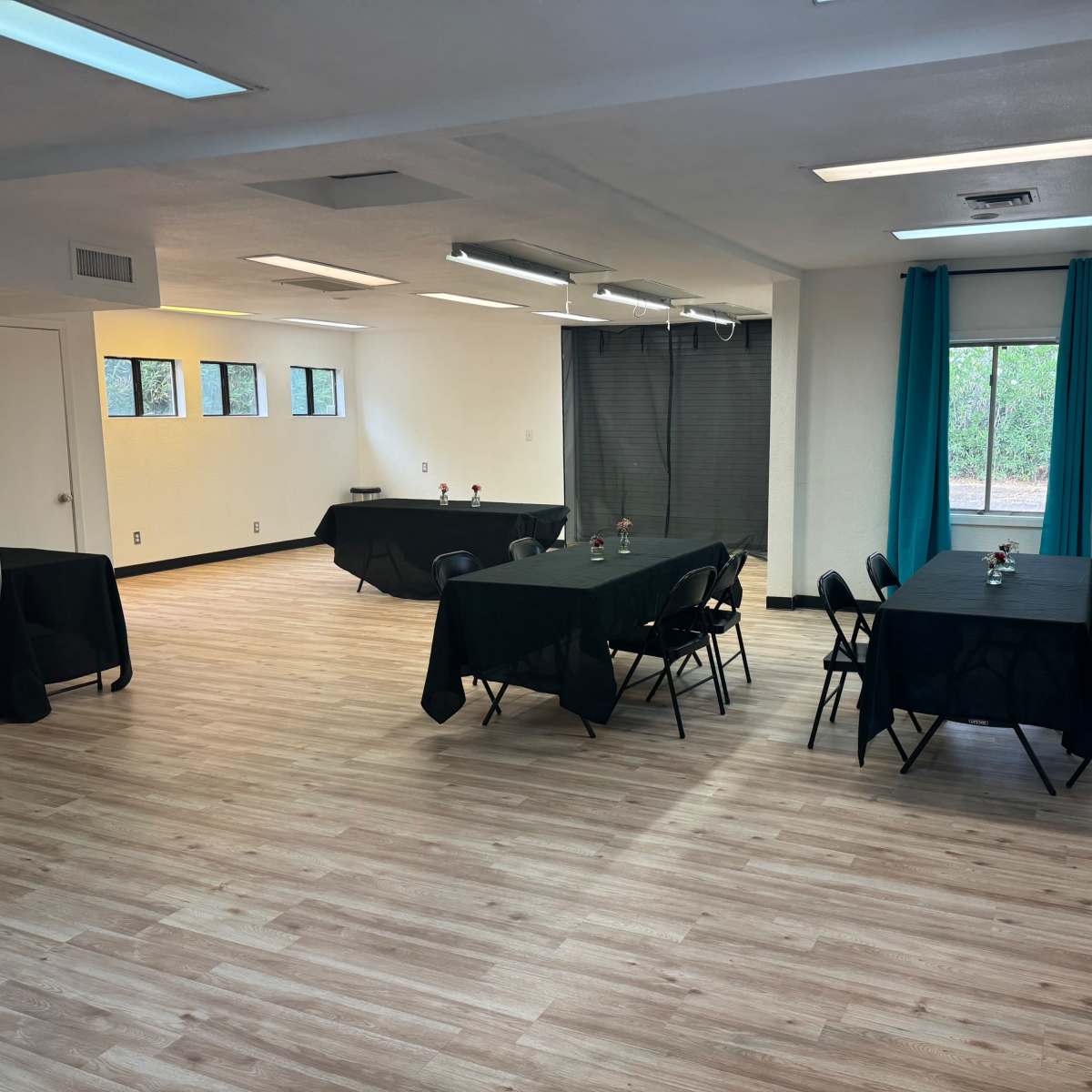 A spacious room with wooden floors, featuring several black tables and chairs arranged with small centerpieces.