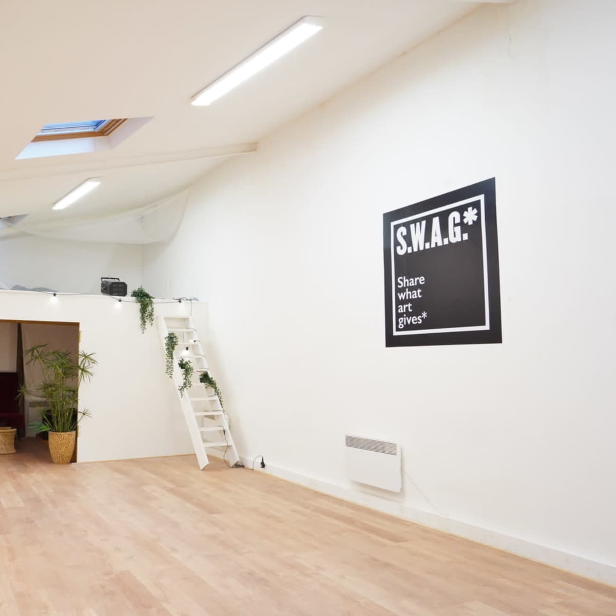 A spacious, well-lit room with wooden flooring, a high ceiling, and minimalist decor, including a wall with the text "S.W.A.G.*" and potted plants.